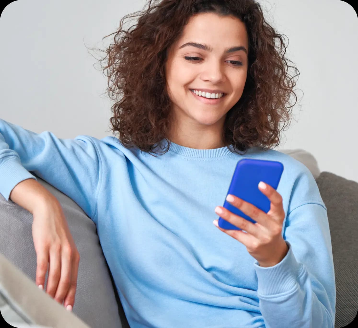 Empreendedora sorri enquanto olha para o celular que está segurando em uma das mãos. Ela tem a pele clara, cabelos ondulados na altura dos ombros e veste uma blusa azul clara. A mulher está sentada em um sofá cinza.