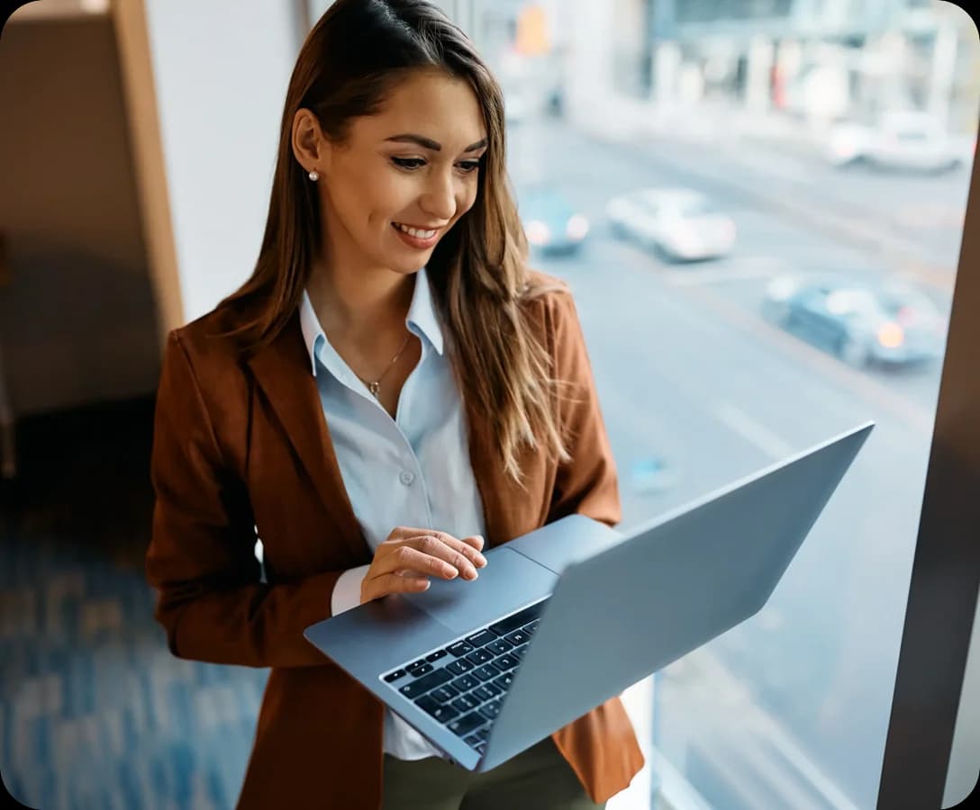 Mulher sorridente, de blazer, usando notebook em um ambiente de escritório com vista para a rua