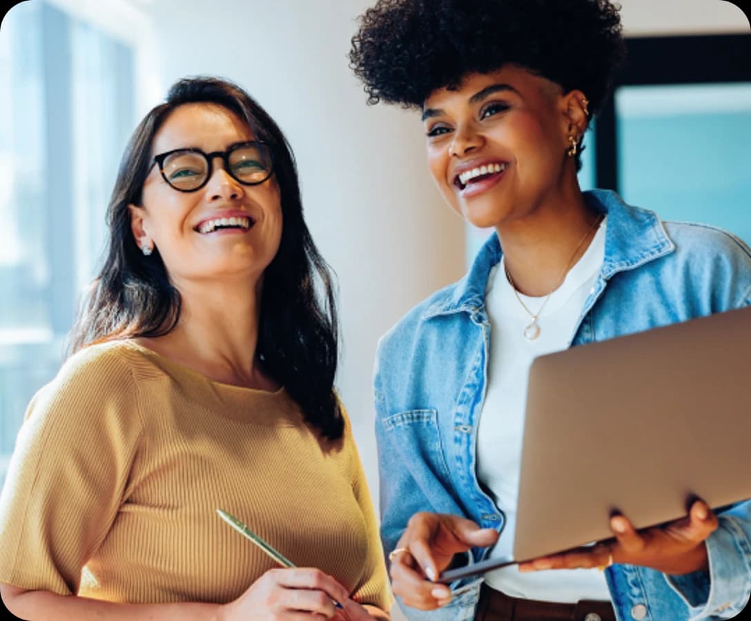 Duas mulheres sorrindo e olhando na mesma direção, uma delas segurando um notebook